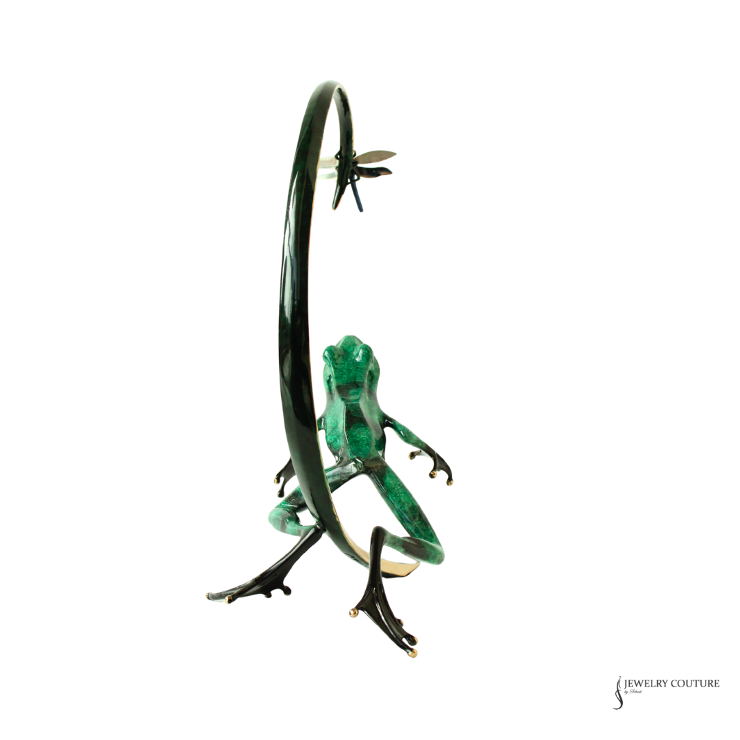 A green frog figurine posed beneath a curved black metal loop, with a small dragonfly above, set against a plain white background.