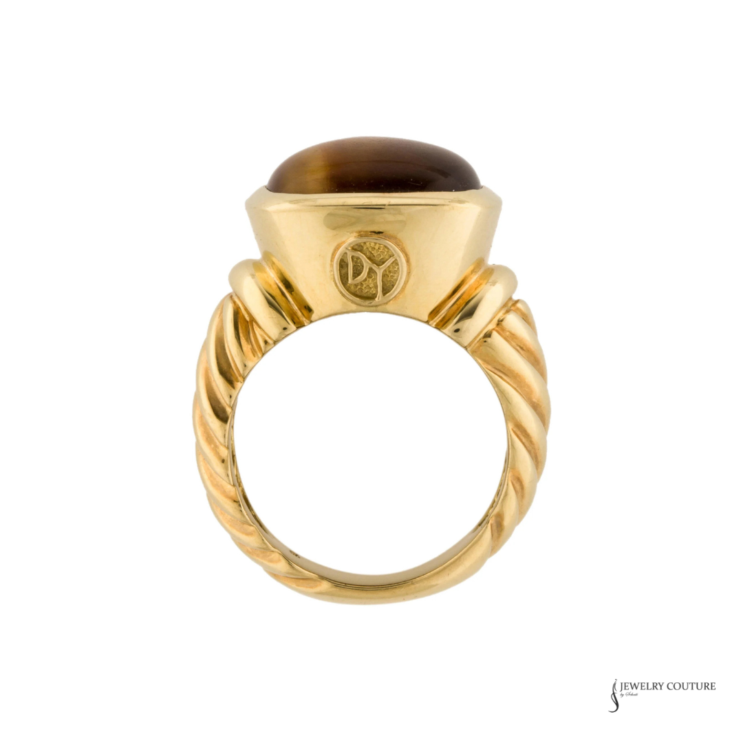 A yellow gold ring featuring a twisted band and a dome-shaped brown tiger's eye stone, shown against a plain white background.
