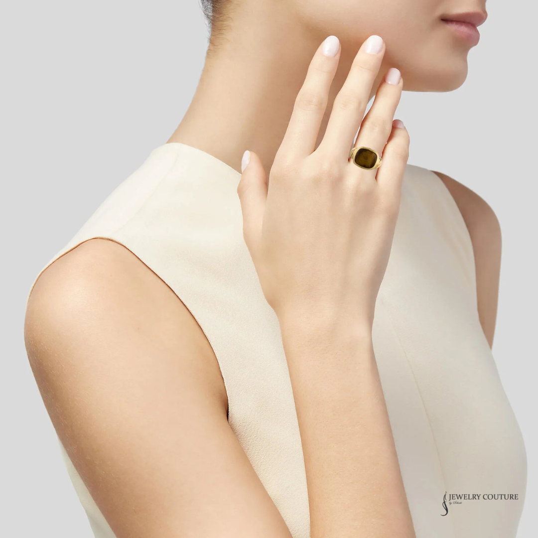 A close-up lifestyle shot of a model wearing a gold signet ring with a dark green stone, styled with a cream-colored sleeveless top against a light gray background.