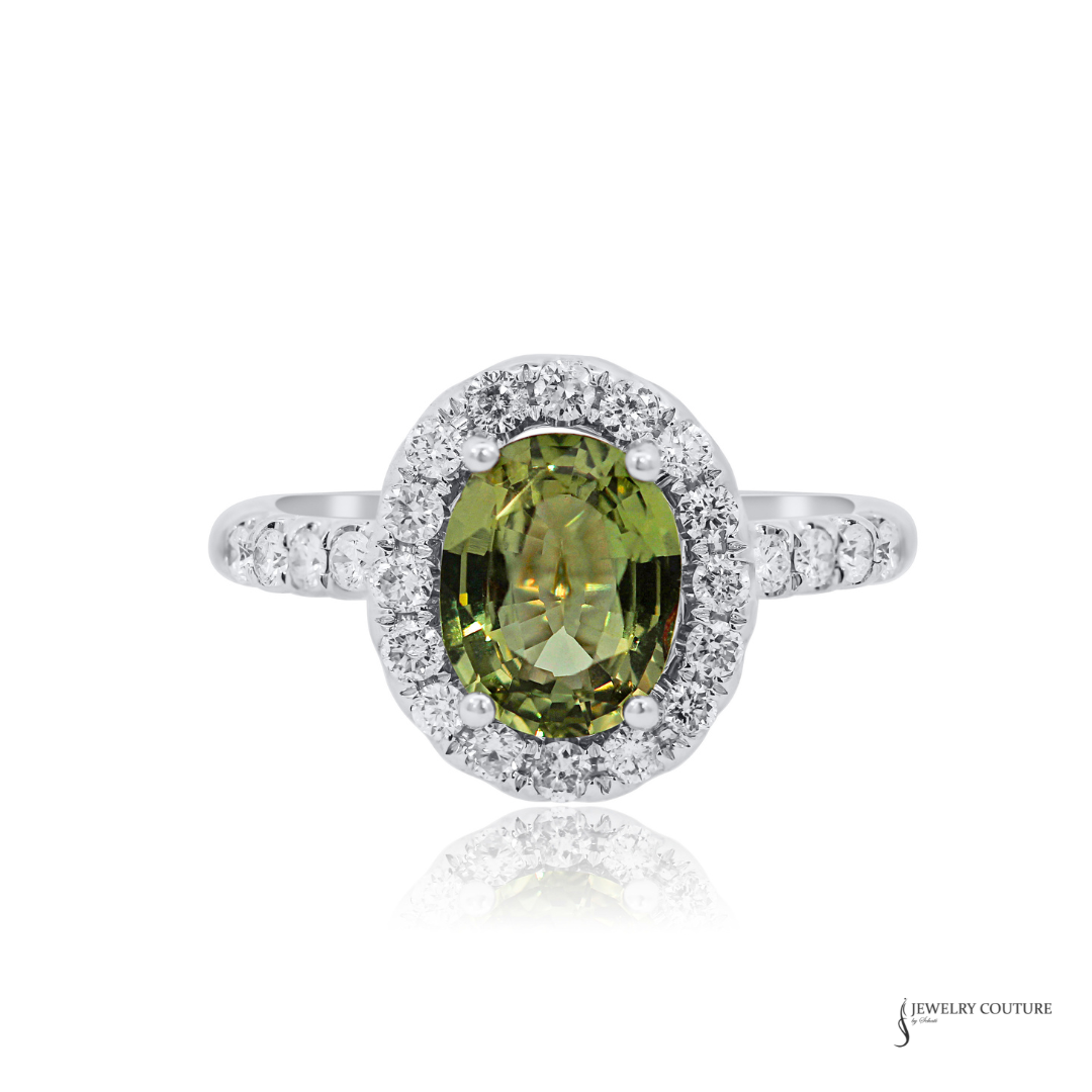 An oval-cut olive green gemstone halo ring in silver-colored metal, featuring a single-prong center stone surrounded by a double halo of small clear stones, with additional clear stones set along the band, shown on a white background with a faint reflection.