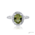 An oval-cut olive green gemstone halo ring in silver-colored metal, featuring a single-prong center stone surrounded by a double halo of small clear stones, with additional clear stones set along the band, shown on a white background with a faint reflection.