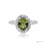 An oval-cut olive green gemstone halo ring in silver-colored metal, featuring a single-prong center stone surrounded by a double halo of small clear stones, with additional clear stones set along the band, shown on a white background with a faint reflection.