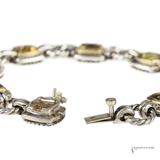 A close-up of a silver-toned chain bracelet with gold-toned bezel-set stones, showing the clasp mechanism and one of the oval links in sharp focus against a white background.