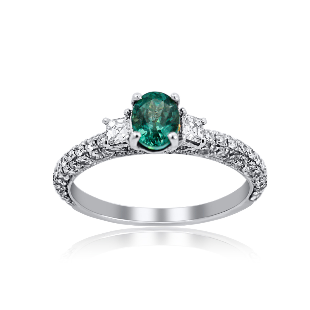 An oval-cut emerald green gemstone ring set in a silver-colored metal band with pavé-set clear accent stones, shown on a white background with a soft reflection below.