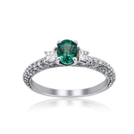 An oval-cut emerald green gemstone ring set in a silver-colored metal band with pavé-set clear accent stones, shown on a white background with a soft reflection below.