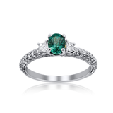 An oval-cut emerald green gemstone ring set in a silver-colored metal band with pavé-set clear accent stones, shown on a white background with a soft reflection below.