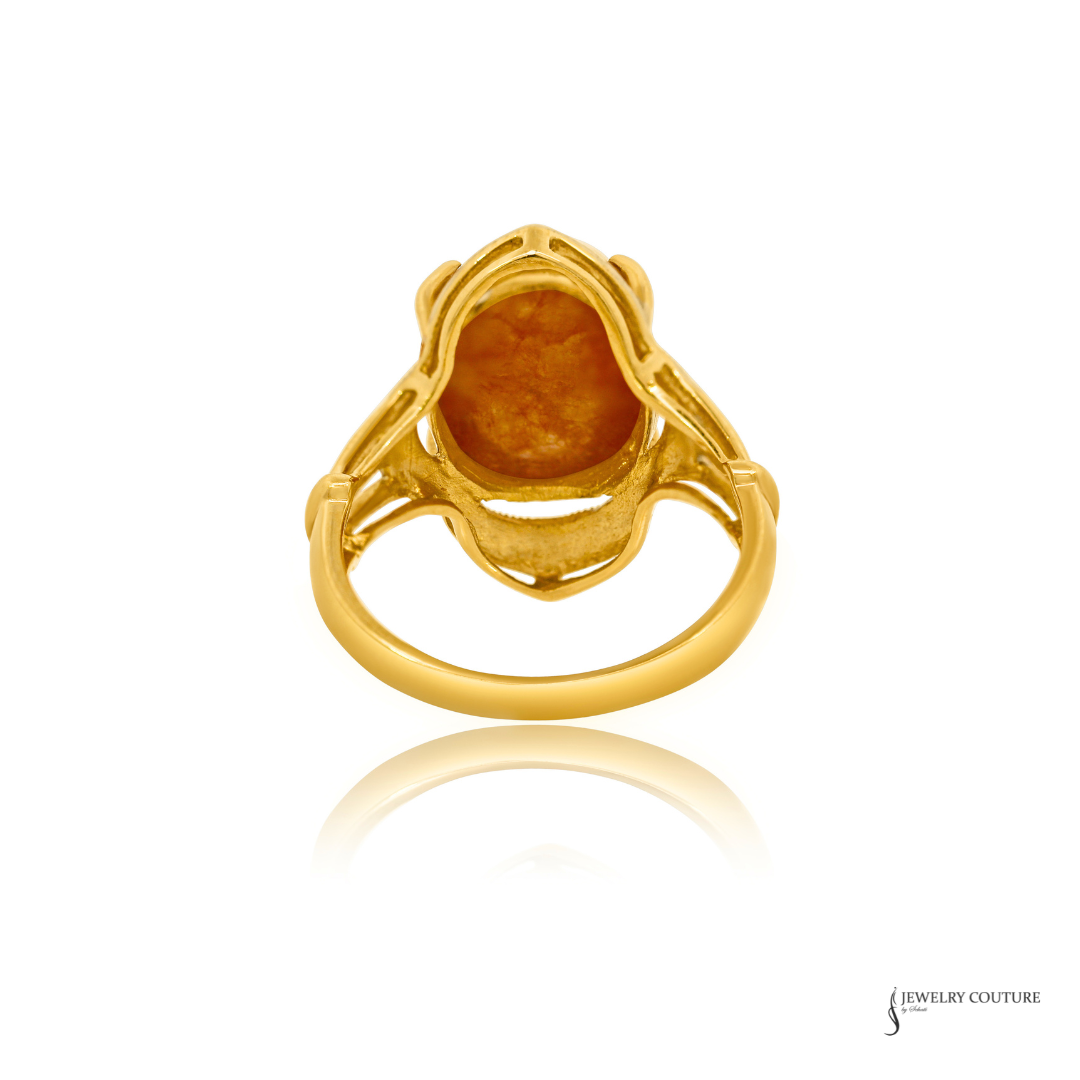 A yellow gold ring featuring a large, irregularly shaped orange-brown gemstone in a decorative setting, shown on a white background with a soft reflection below.