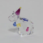 A clear faceted crystal figurine shaped like a cow wearing a purple party hat with a yellow tip, decorated with small multicolored spots, photographed on a plain light gray background.