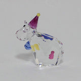 A clear faceted crystal figurine shaped like a cow wearing a purple party hat with a yellow tip, decorated with small multicolored spots, photographed on a plain light gray background.