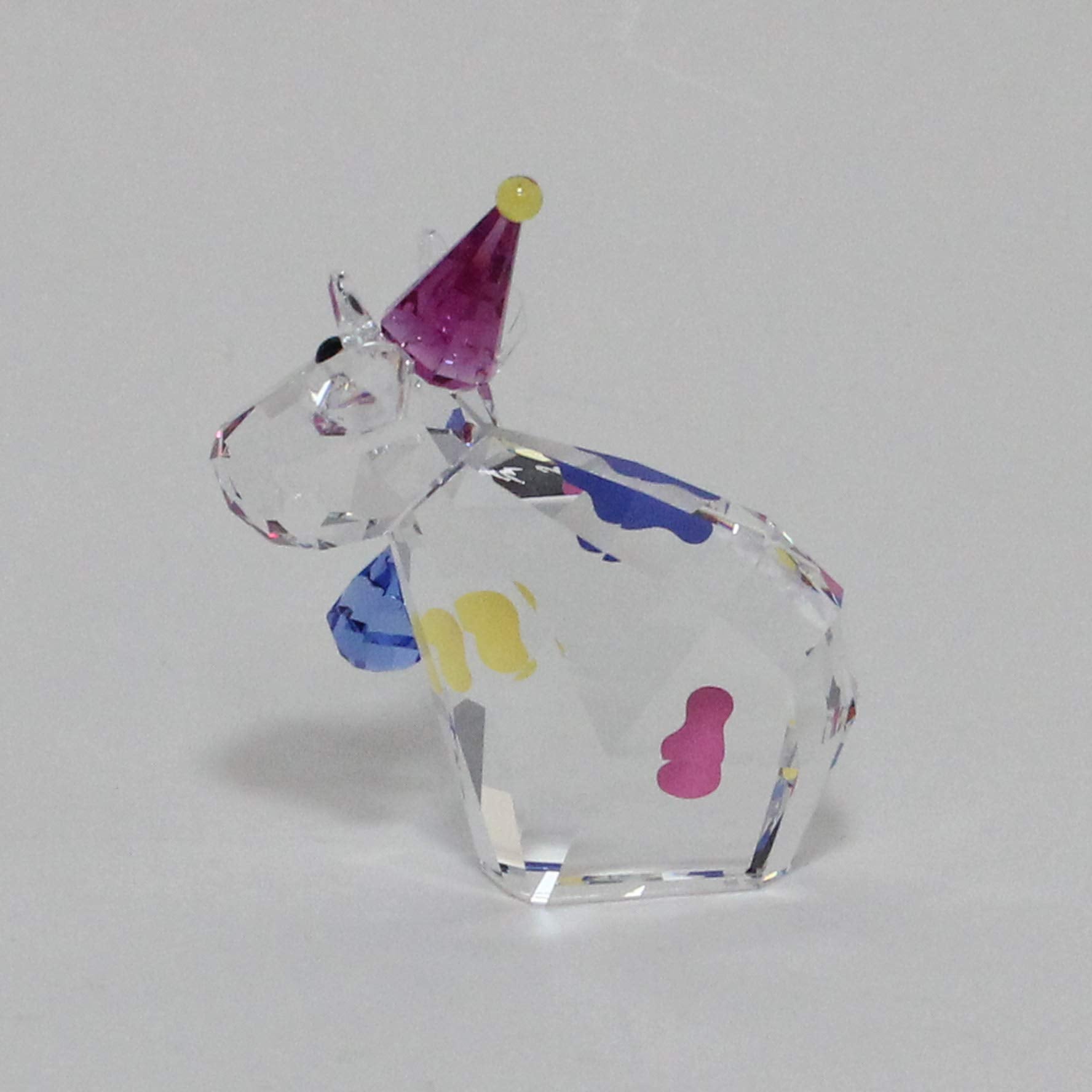 A clear faceted crystal figurine shaped like a cow wearing a purple party hat with a yellow tip, decorated with small multicolored spots, photographed on a plain light gray background.
