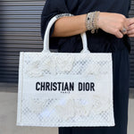 A person wearing a black outfit holds a white Christian Dior tote bag with butterfly embroidery and black lettering, set against a gray metal shutter background.