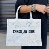 A person wearing a black outfit holds a white Christian Dior tote bag with butterfly embroidery and black lettering, set against a gray metal shutter background.