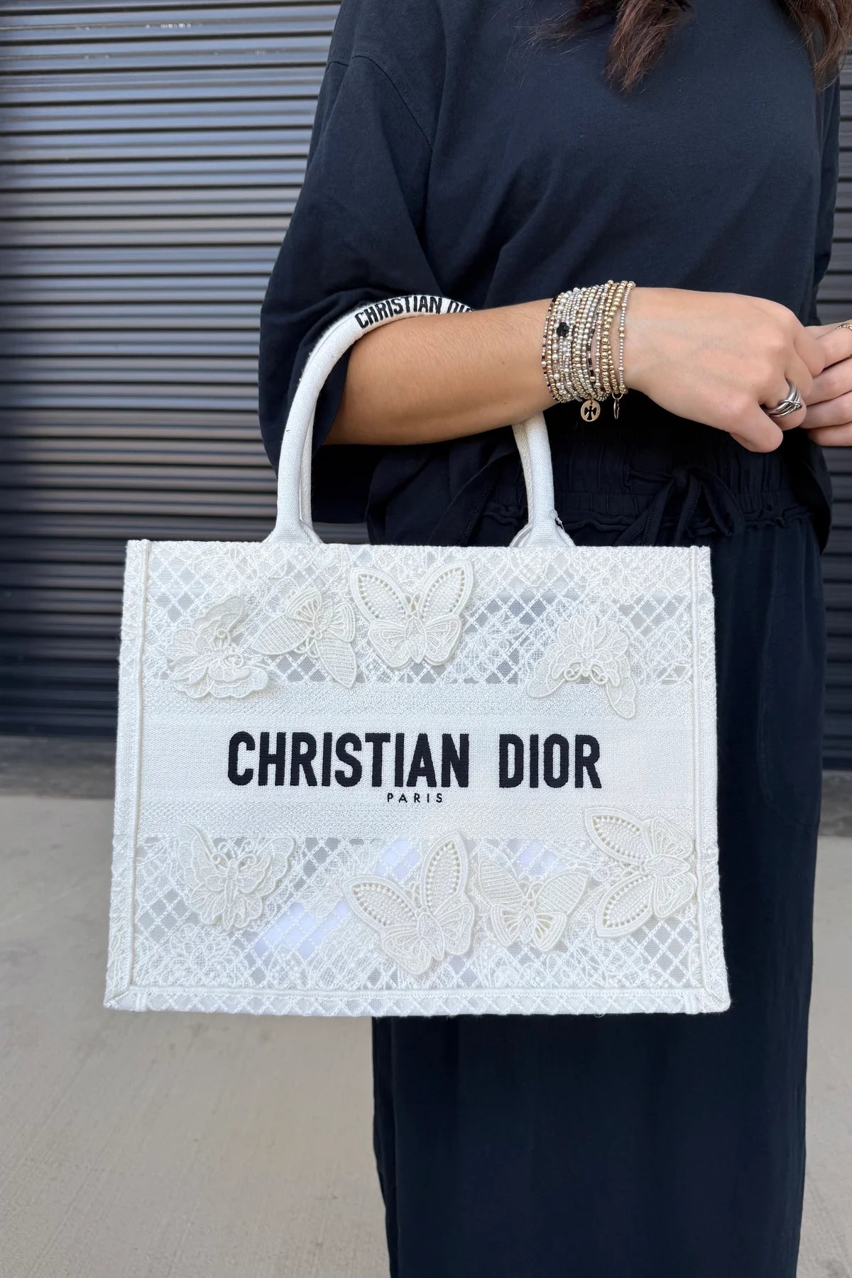 A person wearing a black outfit holds a white Christian Dior tote bag with butterfly embroidery and black lettering, set against a gray metal shutter background.