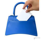 A bright blue leather handbag with a top handle, shown on a white background with a hand inserting a white card into the side slot.