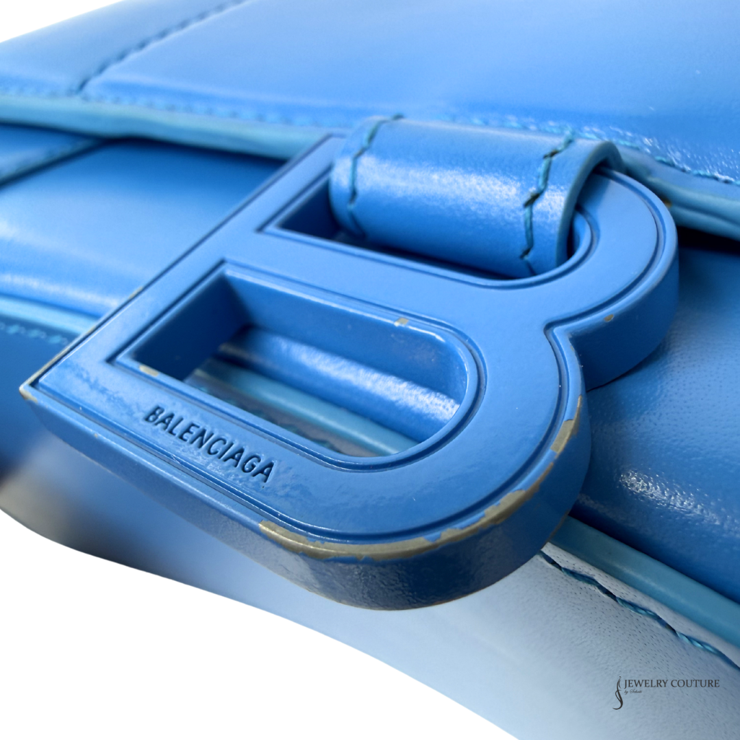 Close-up of a Balenciaga blue leather handbag featuring a large embossed B logo clasp with the brand name 'BALENCIAGA' debossed on it.