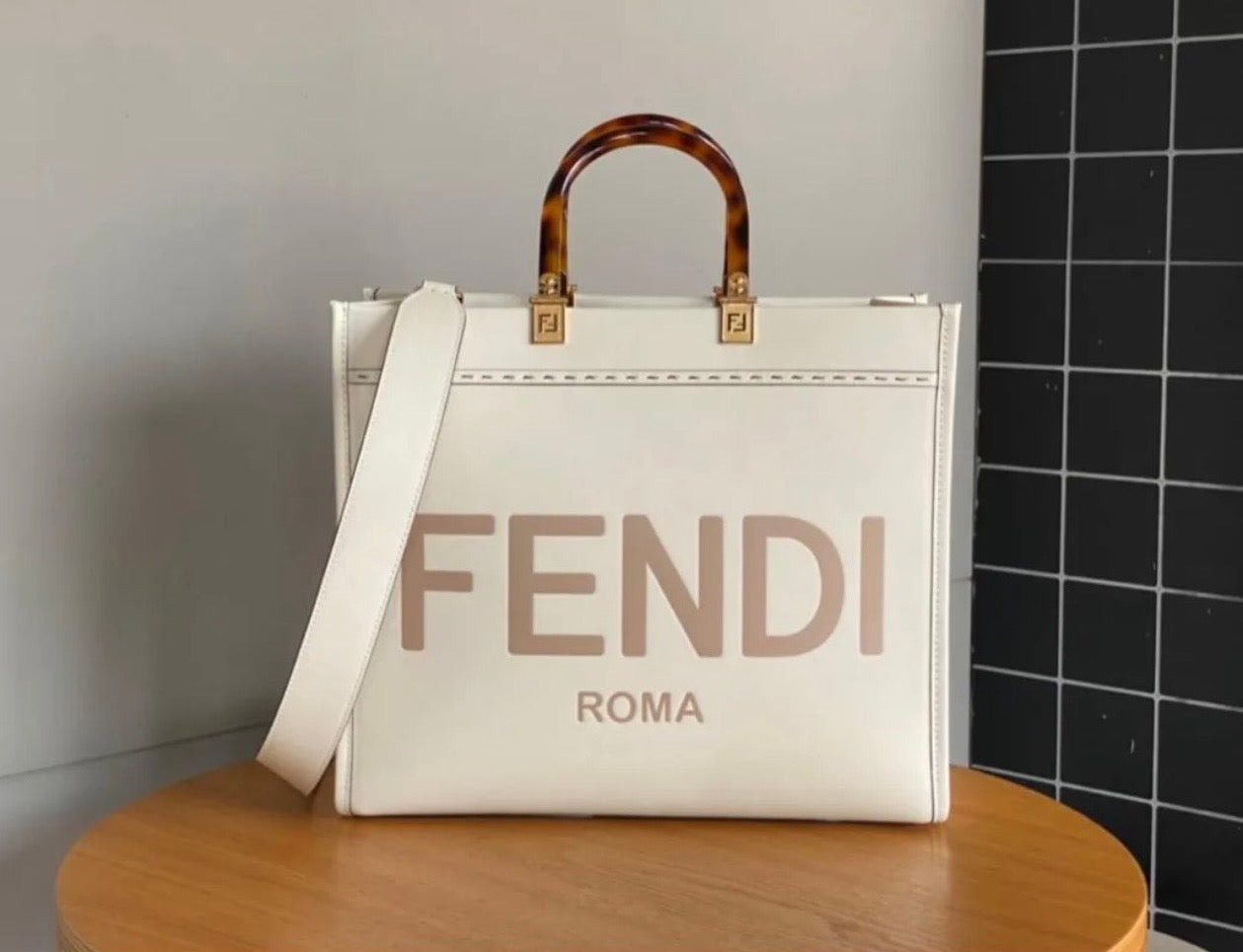 A white Fendi Roma tote bag with a brown faux-bamboo top handle and gold hardware, displayed on a wooden table against a gray wall and black tiled background.