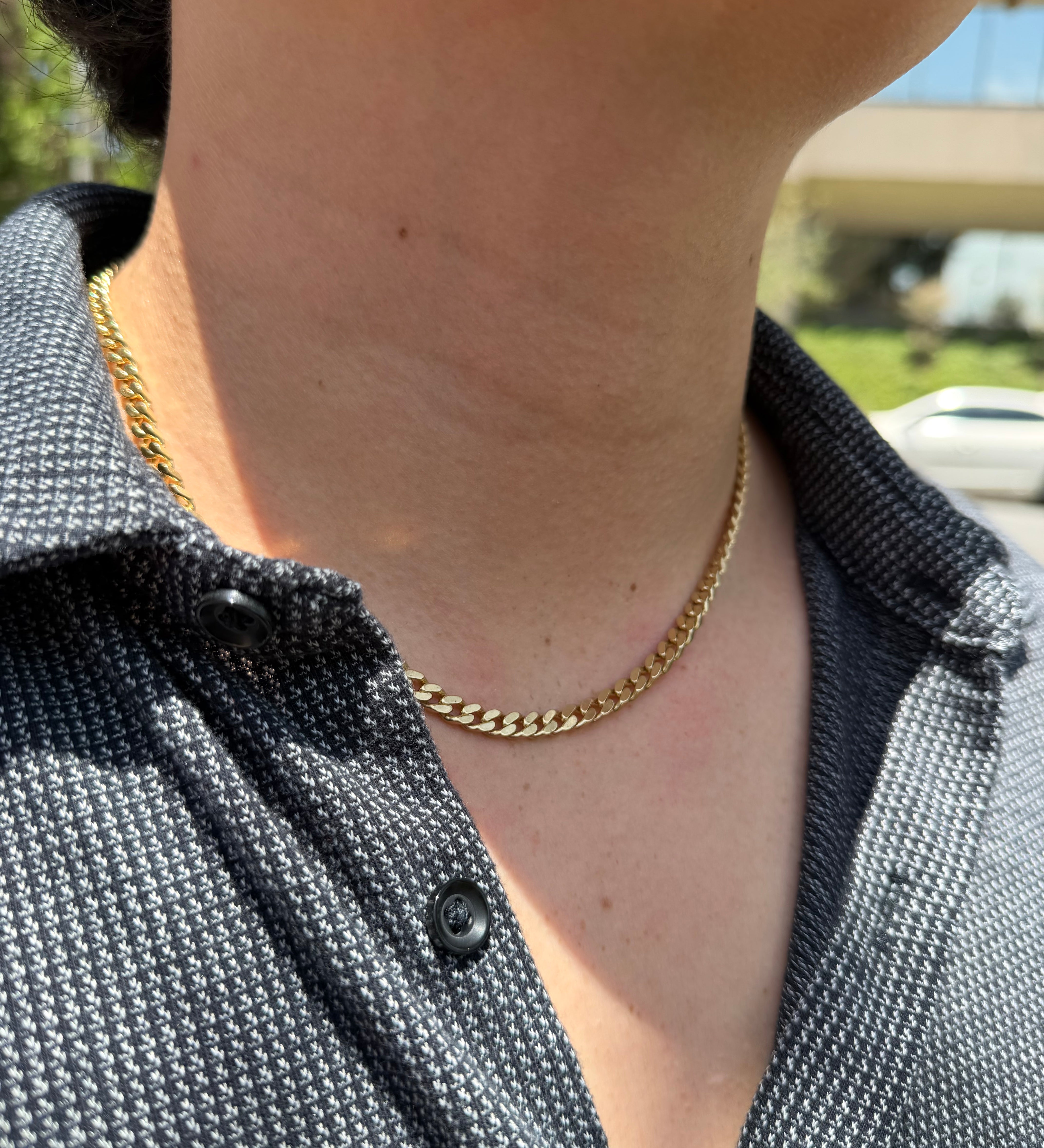 A close-up of a person's neck and chest wearing a gold chain necklace over a dark gray patterned button-up shirt.