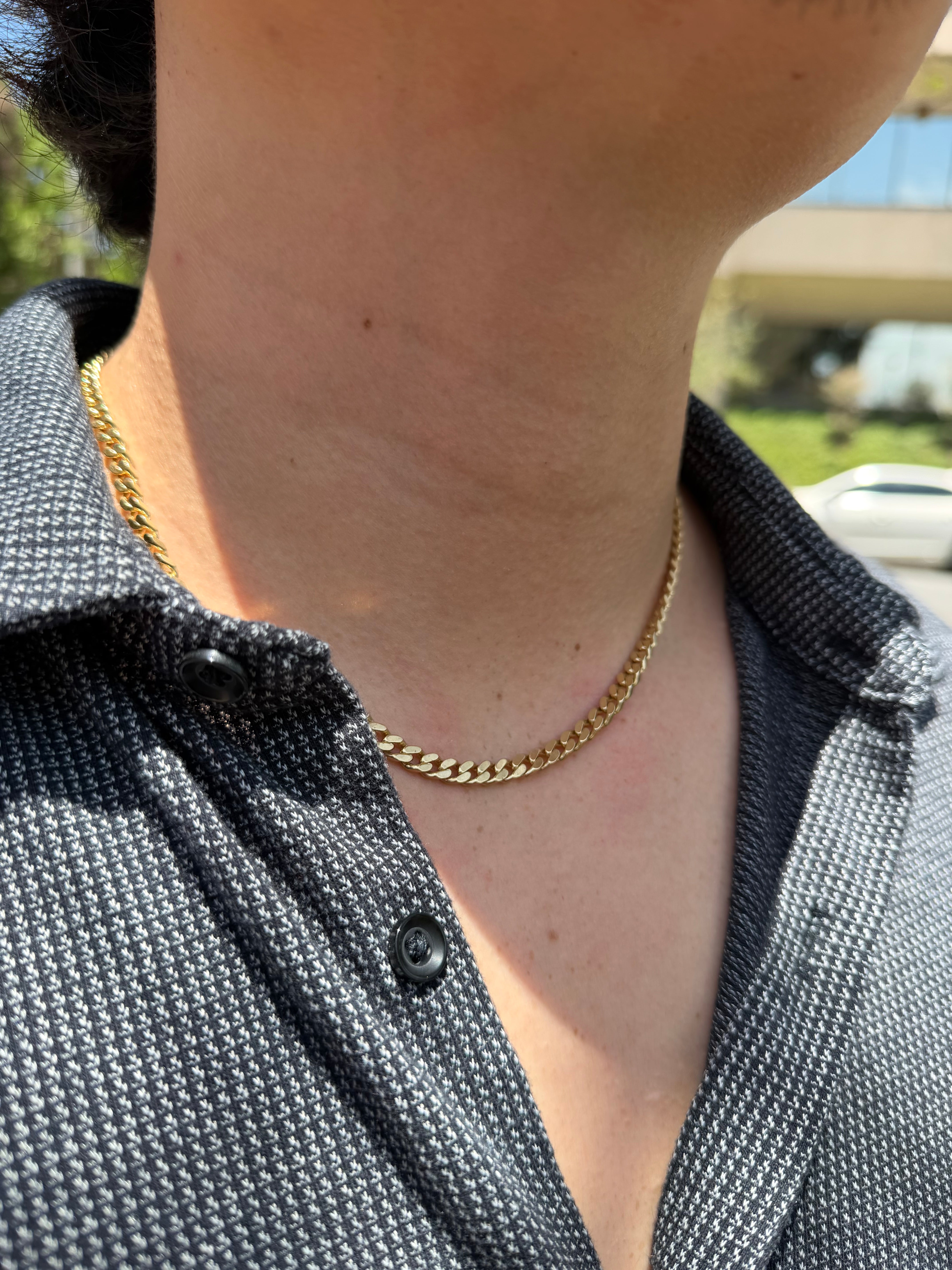 A close-up of a person's neck and chest wearing a gold chain necklace over a dark gray patterned button-up shirt.