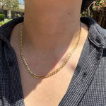 A close-up of a person's neck and chest wearing a gold chain necklace over a dark patterned button-up shirt, with outdoor greenery in the background.