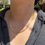 A close-up of a person's neck and chest wearing a gold chain necklace over a dark patterned button-up shirt, with outdoor greenery in the background.
