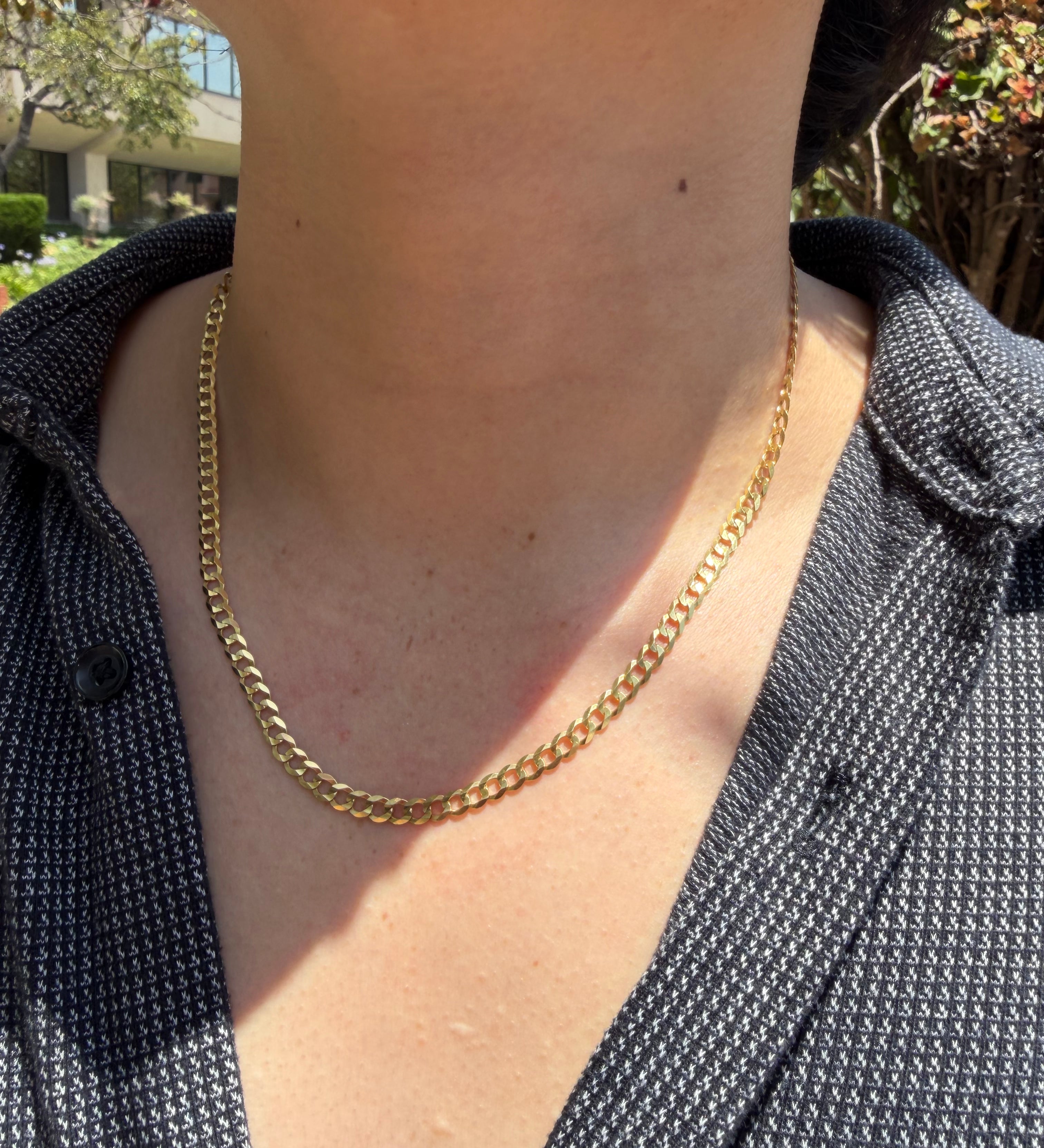 A close-up of a person's neck and chest wearing a gold chain necklace over a dark patterned button-up shirt, with outdoor greenery in the background.