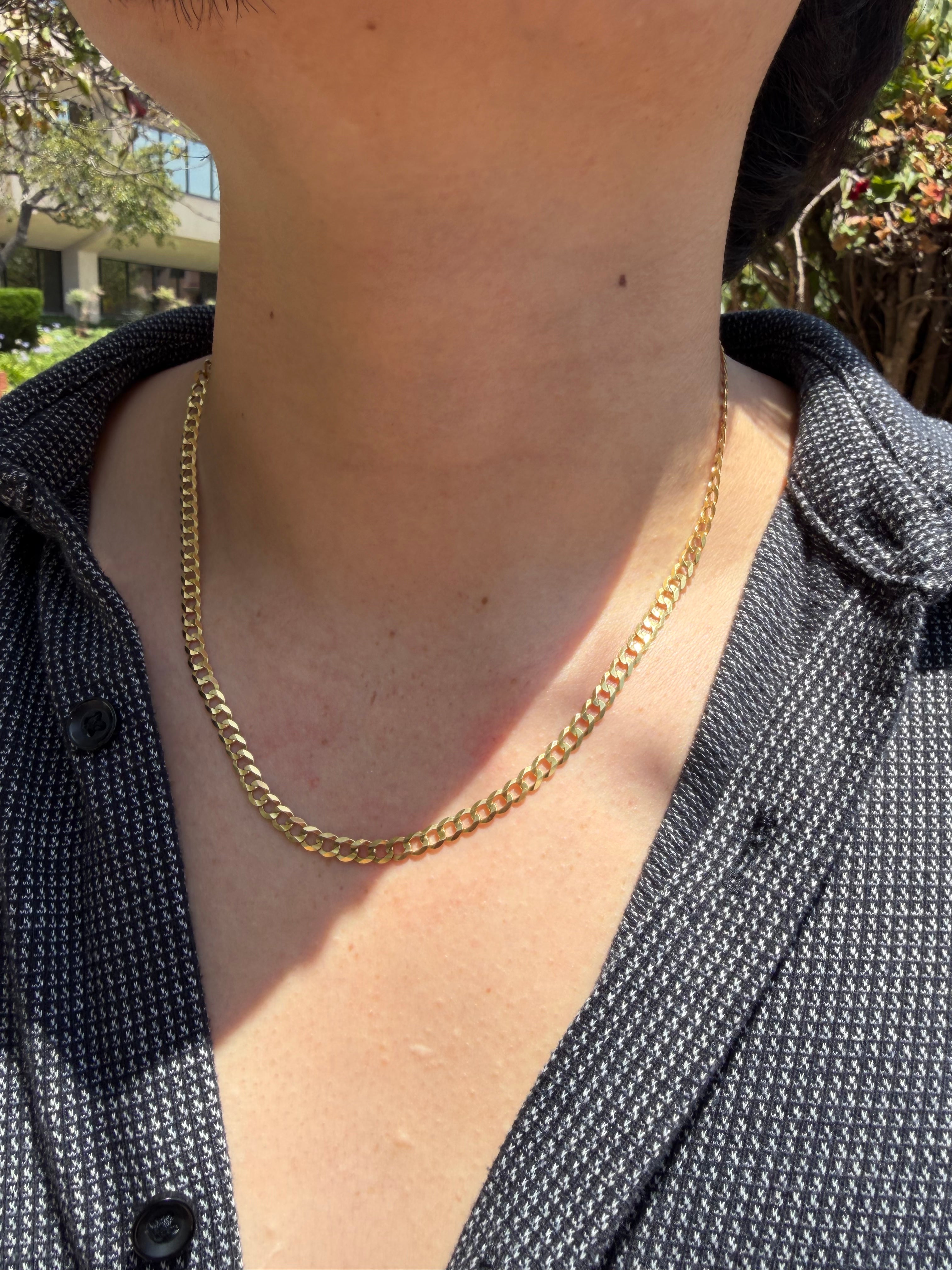 A close-up of a person's neck and chest wearing a gold chain necklace over a dark patterned button-up shirt, with outdoor greenery in the background.