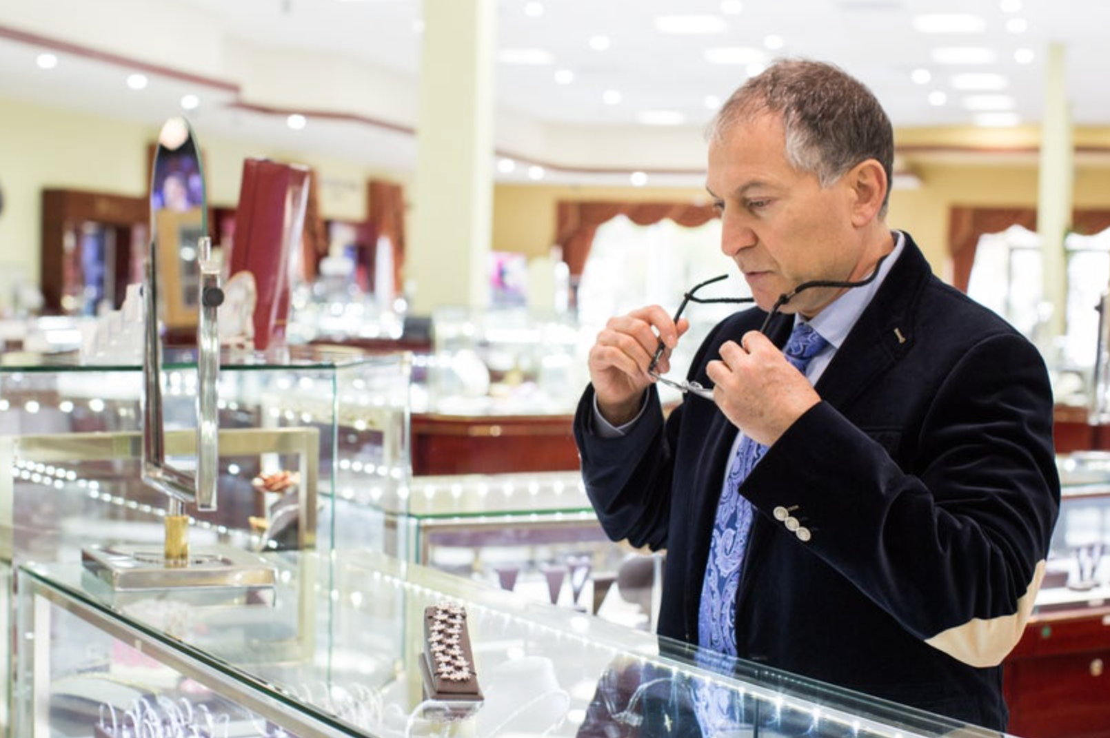 A man in a dark blazer and patterned shirt stands behind a glass jewelry display case in a jewelry store, holding a pair of eyeglasses.