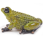 A glossy green glass frog figurine with textured bumpy skin, large black eyes, and black webbed feet with small metallic bronze tips, shown on a plain white background.