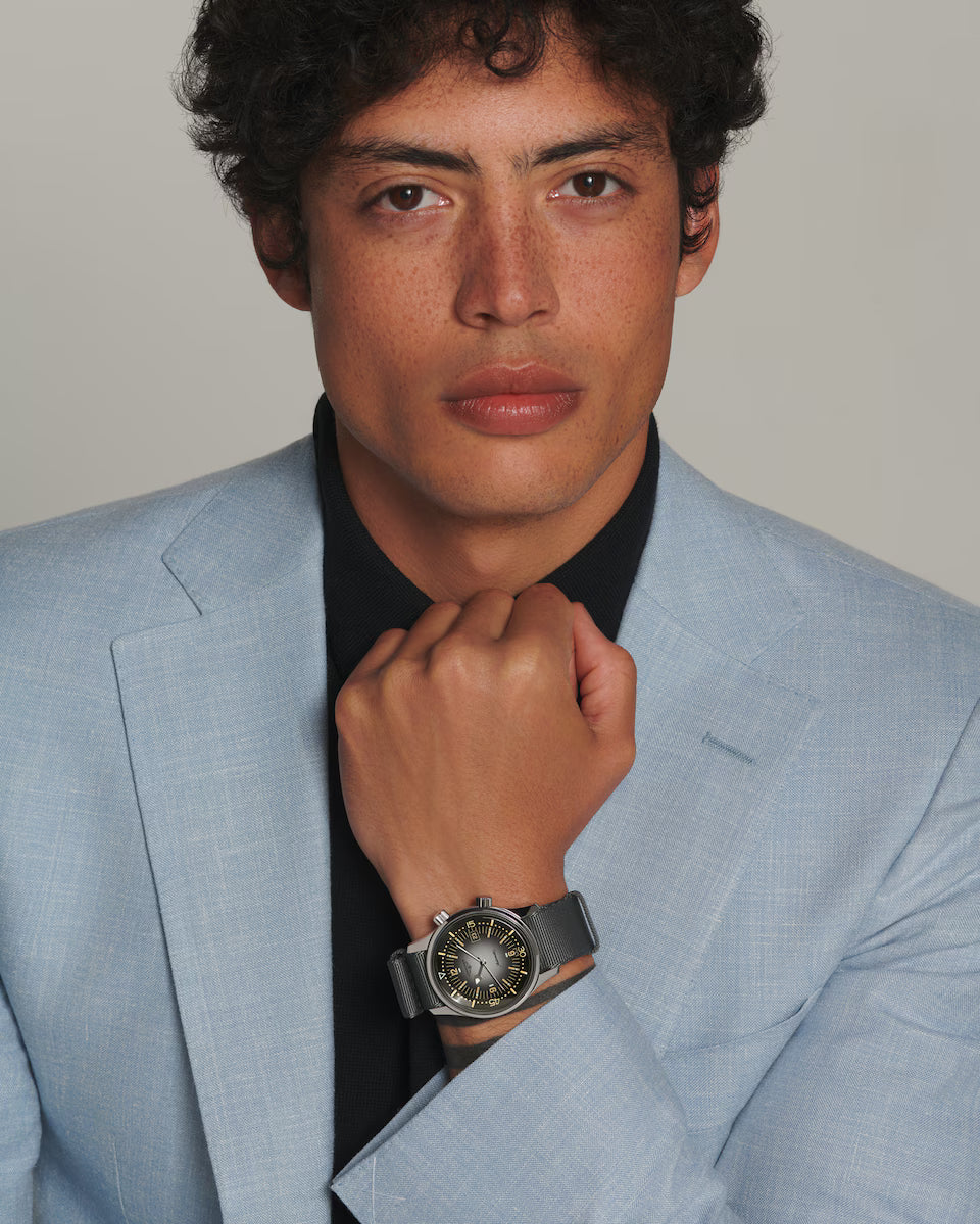 Man wearing a light blue suit with a black shirt and a watch, against a plain background