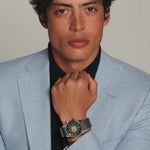 Man wearing a light blue suit with a black shirt and a watch, against a plain background