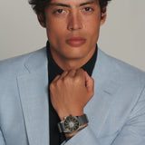 Man wearing a light blue suit with a black shirt and a watch, against a plain background