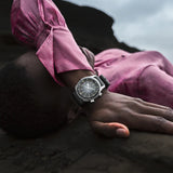 Person wearing a pink shirt and watch, lying on a dark surface with a cloudy sky.