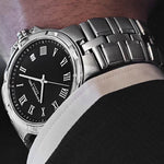 A close-up of a men's stainless steel wristwatch with a black dial and Roman numeral hour markers, worn on a person's wrist with a white dress shirt cuff visible.