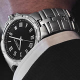 A close-up of a men's stainless steel wristwatch with a black dial and Roman numeral hour markers, worn on a person's wrist with a white dress shirt cuff visible.