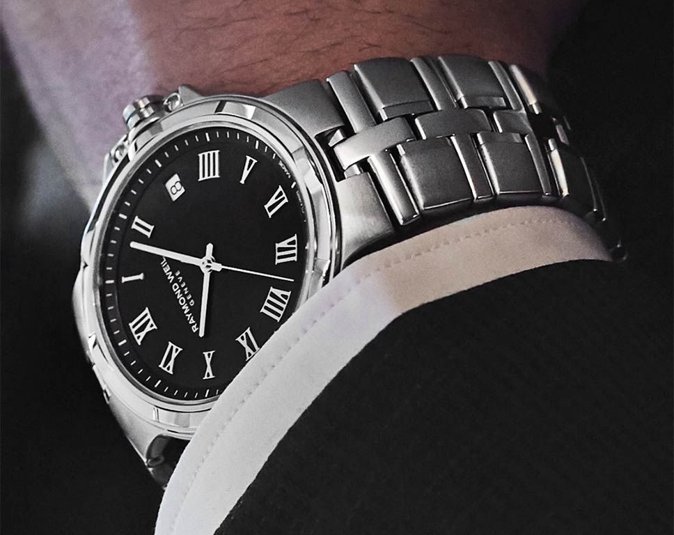 A close-up of a men's stainless steel wristwatch with a black dial and Roman numeral hour markers, worn on a person's wrist with a white dress shirt cuff visible.