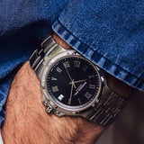 A close-up of a wrist wearing a silver-toned stainless steel watch with a black dial, Roman numeral hour markers, and a date window, partially covered by blue denim.