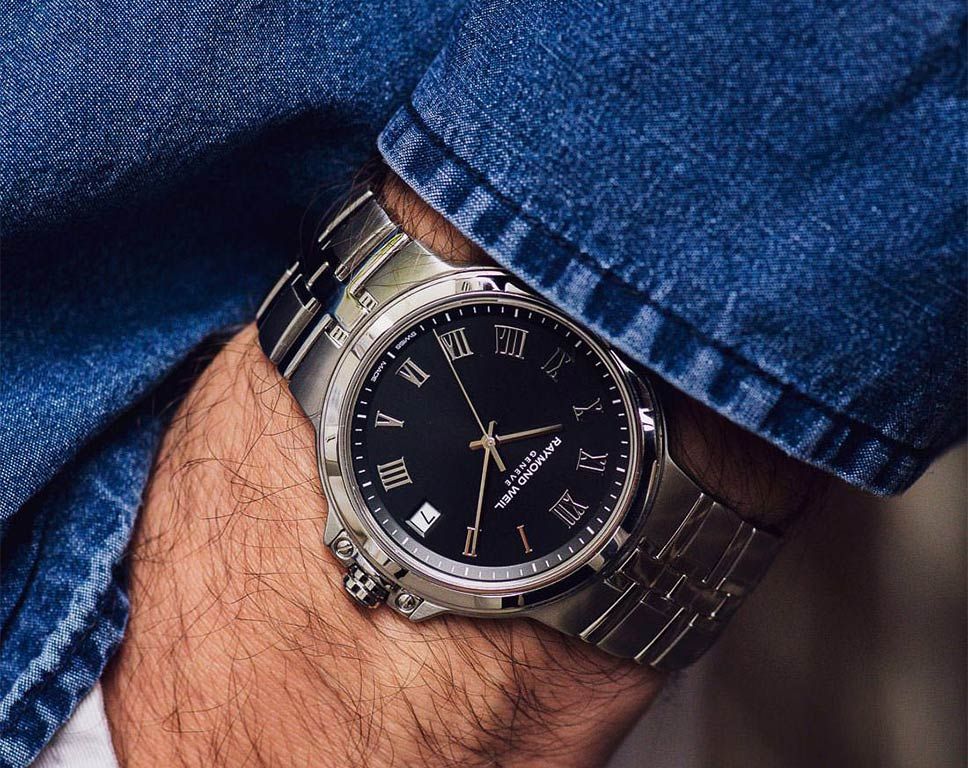 A close-up of a wrist wearing a silver-toned stainless steel watch with a black dial, Roman numeral hour markers, and a date window, partially covered by blue denim.