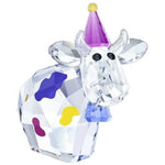 A faceted crystal cow figurine with colorful painted spots in purple, yellow, and blue, wearing a purple party hat with a gold ball topper, isolated on a white background.