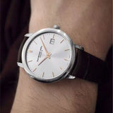 A wristwatch with a silver-toned round case, white dial, and dark brown leather strap worn on a person's wrist.