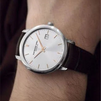 A wristwatch with a silver-toned round case, white dial, and dark brown leather strap worn on a person's wrist.