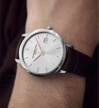A wristwatch with a silver-toned round case, white dial, and dark brown leather strap worn on a person's wrist.