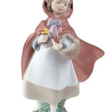 A porcelain figurine of a young girl in a red hooded cloak holding a basket and small yellow flowers, standing on a decorative base against a plain white background.