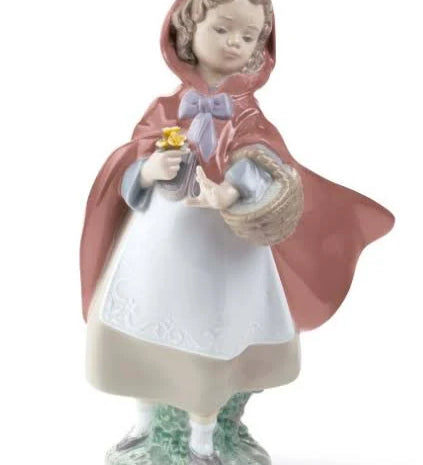 A porcelain figurine of a young girl in a red hooded cloak holding a basket and small yellow flowers, standing on a decorative base against a plain white background.
