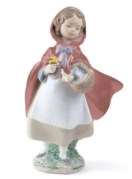 A porcelain figurine of a young girl in a red hooded cloak holding a basket and small yellow flowers, standing on a decorative base against a plain white background.
