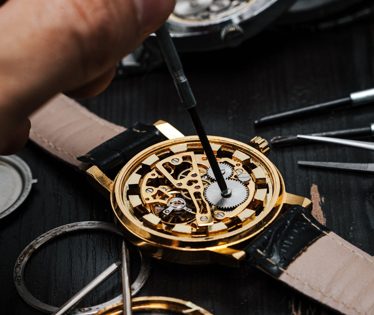 Person working on a gold watch with visible mechanical components on a dark surface.