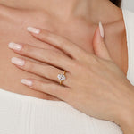 A close-up of a woman's hand resting on her chest, wearing a yellow gold ring with a clear round-cut gemstone. The background shows her skin, white top, and manicured nails.