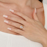 A close-up of a woman's hand resting on her chest, wearing a yellow gold ring with a clear round-cut gemstone. The background shows her skin, white top, and manicured nails.
