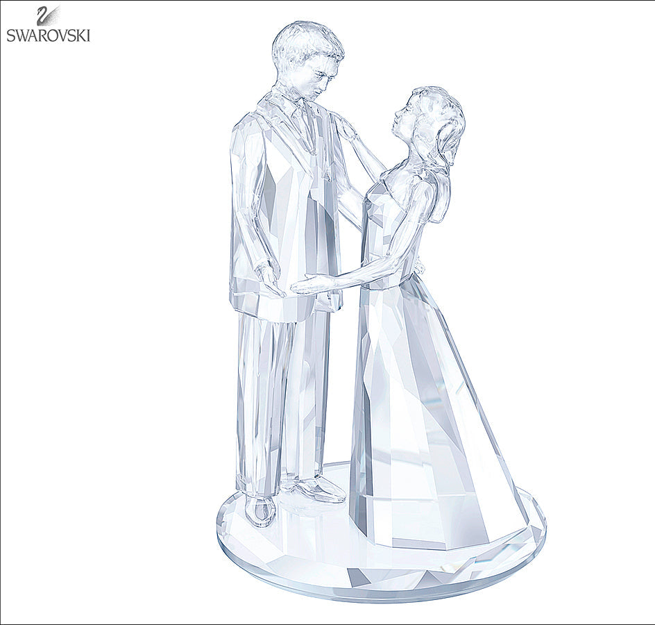 A clear crystal figurine of a groom and bride holding hands, standing on a round base. The Swarovski brand logo is visible in the top-left corner.