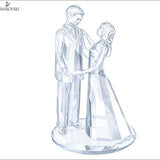 A clear crystal figurine of a groom and bride holding hands, standing on a round base. The Swarovski brand logo is visible in the top-left corner.