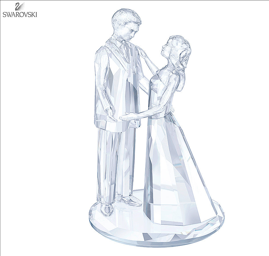 A clear crystal figurine of a groom and bride holding hands, standing on a round base. The Swarovski brand logo is visible in the top-left corner.